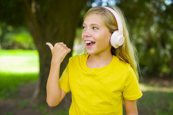 Kaukasisches kleines Mädchen in gelbem T-Shirt, das draußen steht, hört Audio-Track über drahtlose Kopfhörer zeigt Daumen weg Werbung für Kopierraum - Foto, Bild