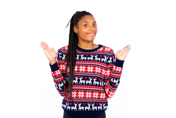 Belki evet ya da hayır. Beyaz duvara karşı Noel kazağı giyen şüpheli Afrikalı Amerikalı kadın şaşkınlık içinde omuz silkiyor, ne istediğine karar vermeye çalışıyor. Tereddüt ve belirsizlik. - Fotoğraf, Görsel