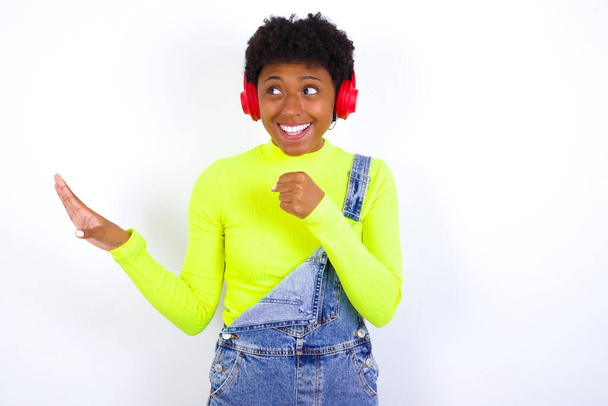 Счастливая молодая афроамериканская женщина с короткими волосами носить джинсы в целом против белой стены поет любимую песню держит руку у рта, как будто микрофон носит беспроводные наушники, слушает музыку - Фото, изображение