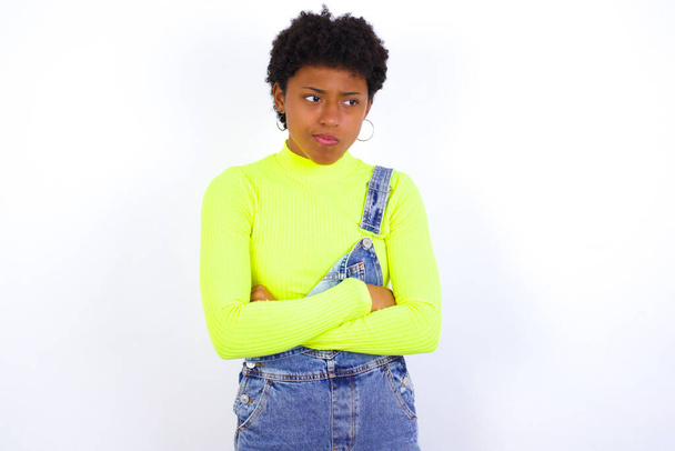 Δυσαρεστημένη νεαρή Αφρο-Αμερικανίδα με κοντά μαλλιά που φοράει τζιν συνολικά ενάντια στο λευκό τοίχο με κακή συμπεριφορά, τα χέρια σταυρωμένα κοιτάζοντας πλάγια. Αρνητικά συναισθήματα ανθρώπινης έκφρασης προσώπου. - Φωτογραφία, εικόνα