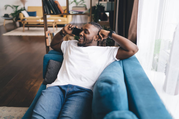 Happy African American bearded male in casual clothes sitting with back on cozy sofa in spacious living room and talking on smartphone while making hand gesture - Fotoğraf, Görsel