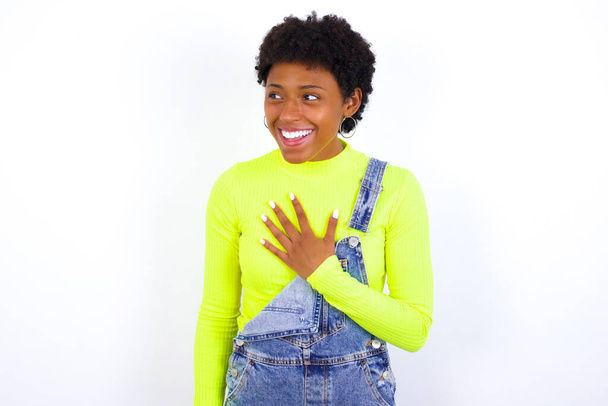 Joven mujer afroamericana alegre con el pelo corto usando denim en general contra la pared blanca expresa emociones positivas recuerda algo divertido mantiene la mano en el pecho y se ríe felizmente. - Foto, imagen