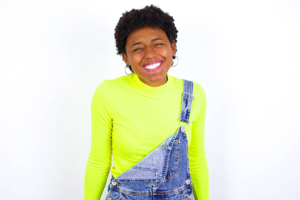 Θετική νεαρή Αφροαμερικανή γυναίκα με κοντά μαλλιά φορώντας τζιν συνολικά κατά λευκό τοίχο με υπερευχαριστημένη έκφραση κλείνει τα μάτια και γελάει δείχνει λευκά τέλεια δόντια - Φωτογραφία, εικόνα
