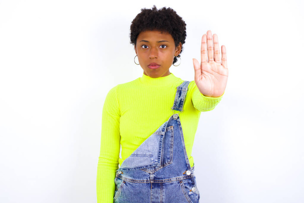 joven mujer afroamericana con el pelo corto con denim en general contra la pared blanca haciendo gesto de parada con la palma de la mano. Expresión de advertencia con gesto negativo y serio en la cara. - Foto, imagen