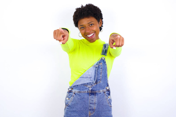 Κοντινό πλάνο πορτραίτο μιας έκπληκτης νεαρής Αφροαμερικανίδας με κοντά μαλλιά που φοράει τζιν συνολικά στον λευκό τοίχο δείχνοντας με δύο δάχτυλα την κάμερα λέγοντας: Διαλέγω εσένα!, κοιτώντας ψηλά με ανοιχτό στόμα. - Φωτογραφία, εικόνα
