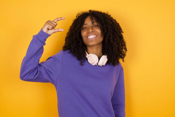 портрет молодої виразної афроамериканки з навушниками на жовтому фоні, що показує жест малого розміру - Фото, зображення