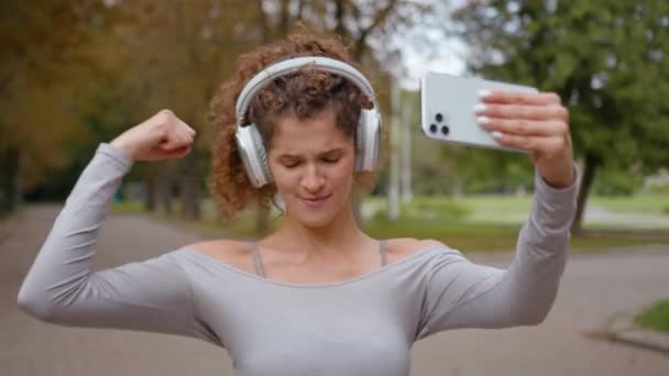 Кавказская сильная спортивная девушка, демонстрирующая чемпионский жест, изгибает бицепсы, показывая на камере запись звонка мобильного телефона с тренировкой в городе с наушниками на открытом воздухе - Кадры, видео