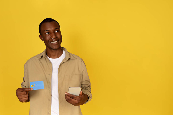 Усміхнений пенсіонерський чорний хлопець використовує телефон і кредитну картку, мрію, думати, ізольований жовтий студійний фон. Реклама та пропозиція, додаток для банківської справи та продажу, онлайн-покупки, оплата грошей - Фото, зображення
