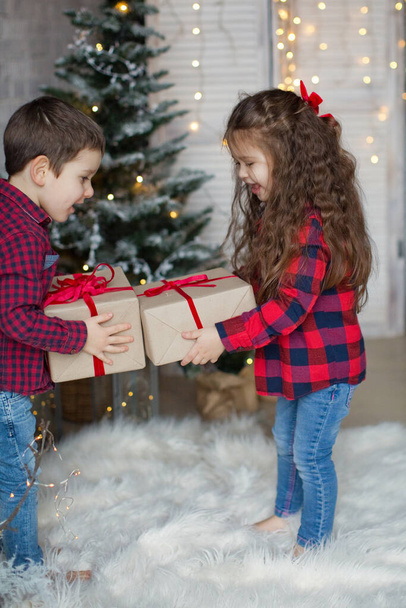 Mały chłopiec i dziewczynka w czerwonych koszulach trzymać pudełka prezentów w pobliżu choinki w jasnym wnętrzu Bożego Narodzenia. Słodkie dzieci Boże Narodzenie - Zdjęcie, obraz