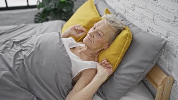 Donna anziana fiduciosa con i capelli grigi, godendo di una mattinata rilassante svegliarsi nel comfort della sua camera da letto, tenendo uno smartphone mentre disteso su un letto accogliente, con un sorriso raggiante. - Filmati, video