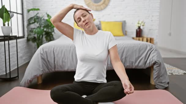mooie jonge blonde vrouw stretching haar nek op slaapkamer vloer, wakker met een glimlach, ondergedompeld in ontspannende yoga - Video