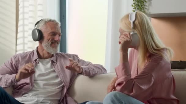 Joyful idosos caucasiano casado casal avós homem mulher ouvir música usando fones de ouvido dançando brincando em torno de se divertir despreocupado avó avô aproveitando fim de semana em casa sofá - Filmagem, Vídeo