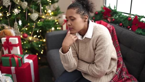 Αφροαμερικανή γυναίκα που βαριέται να κάθεται δίπλα στο χριστουγεννιάτικο δέντρο στο σπίτι - Πλάνα, βίντεο