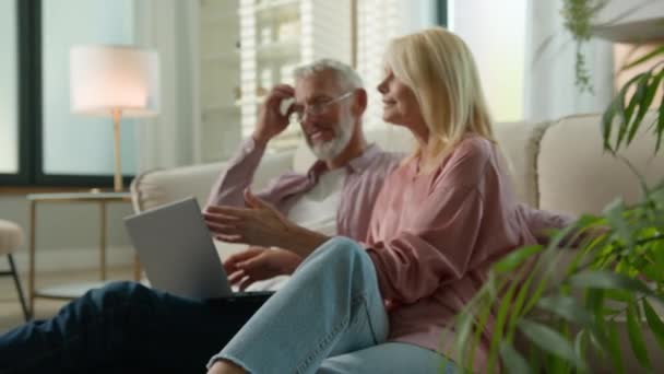 Heureux retraité sans soucis vieux couple marié senior homme femme grand-père parler rire détente ensemble à la maison canapé faire l'ordre dans la boutique en ligne navigation sur les médias sociaux à l'aide d'un ordinateur portable - Séquence, vidéo