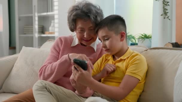 Любящий кавказский внук мальчик ребенок ребенка обучения пожилой женщины бабушка использовать смартфон мобильного приложения покупки в интернет-магазине просматривать социальные сети два поколения семьи проводит время вместе на дому - Кадры, видео