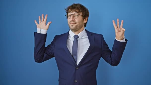 Веселий молодий чоловік у діловому костюмі посміхається і вказує, показуючи номер вісім з пальцями. ізольовані на синьому фоні. - Кадри, відео