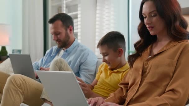 Függője a modern technológia apa anya fia használ különböző szerkentyűk boldog kaukázusi család időt töltenek együtt szülők dolgozik távoli laptop otthon kanapén gyerek fiú gyermek videojáték telefonon - Felvétel, videó