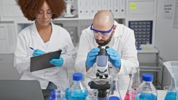 Inside the buzzing lab, two committed scientists, man and woman, analysis partners, seated together, gloved hands busy writing on clipboard, eyes glued to the microscope in serious research - Footage, Video