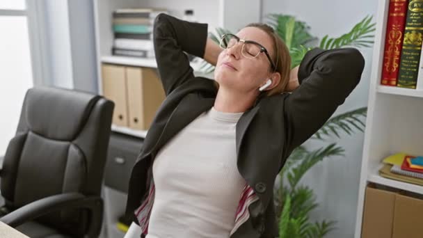 Rilassato giovane lavoratrice bionda, mani sulla testa, godendo la sua musica al lavoro in ufficio interno - Filmati, video