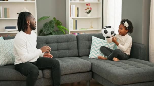 Африканский американец отец и дочь улыбаются уверенно играя на диване, передавая футбольный мяч дома - Кадры, видео