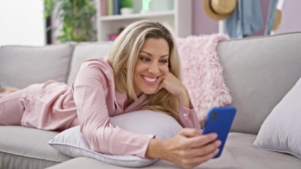 Nuori blondi nainen, jolla on videopuhelu makaa sohvalla kotona - Materiaali, video