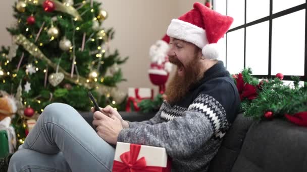 hombre feliz en un sombrero de Santa Claus sentado en el sofá y charlando en un teléfono inteligente - Metraje, vídeo