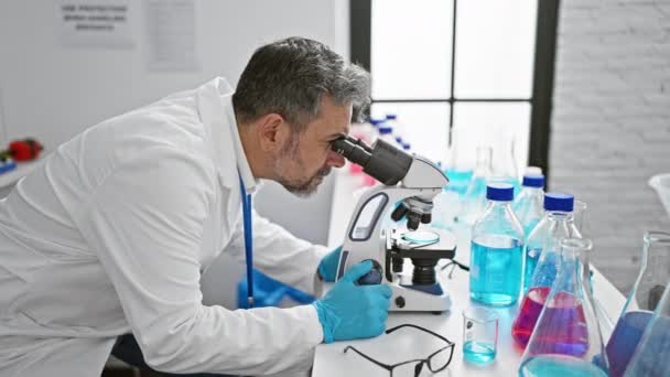 Sebevědomý mladý hispánský vědec s šedivými vlasy, usmívá se, když pracuje s mikroskopem v laboratoři, dělá lékařské objevy - Záběry, video