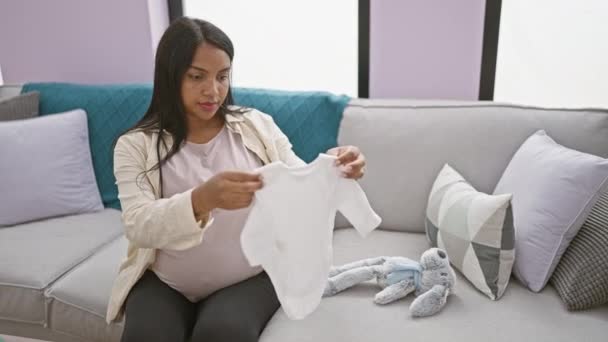 Mujer embarazada joven radiante plegando alegremente la ropa del bebé tocando su vientre, relajándose en un cómodo sofá en casa - Metraje, vídeo