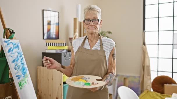 Grauhaarige Seniorin mit Schürze und Brille steht zweifelnd an ihrer Staffelei, Pinsel und Palette in der Hand und denkt über ihren nächsten Schlaganfall im Herzen ihres Ateliers nach.. - Filmmaterial, Video