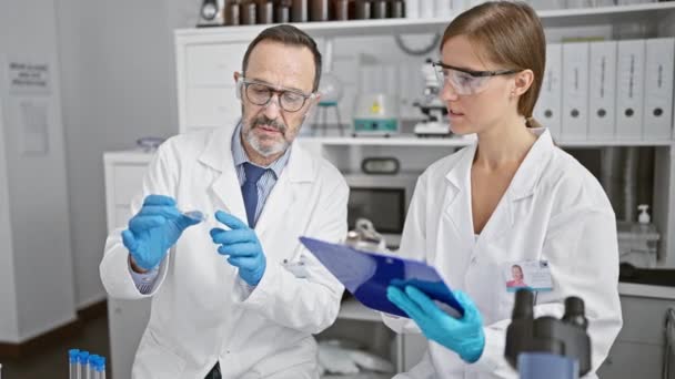 Deux scientifiques dévoués en blouse de laboratoire, travaillant diligemment ensemble au cœur de leur laboratoire, blottis sur un précieux échantillon, prenant studieusement des notes sur le presse-papiers. - Séquence, vidéo