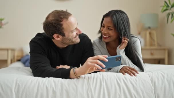 Kaunis pari makaa sängyssä katsomassa videota älypuhelimella makuuhuoneessa - Materiaali, video
