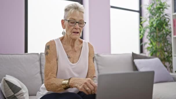 Mujer mayor de cabello gris grave, enfocada en usar un portátil, sentada en un sofá en casa, tocando el estómago con un dolor, signo de enfermedad o enfermedad - Imágenes, Vídeo