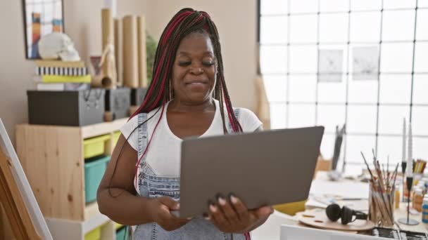 Magabiztos afro-amerikai női művész copfokkal, mosolyog, miközben vászonra fest, és laptopot használ egy hangulatos művészeti stúdióban. - Felvétel, videó