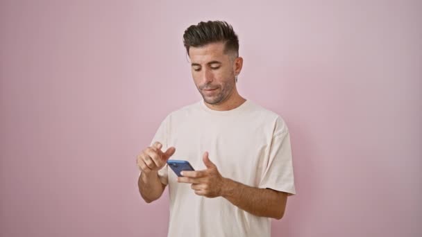 Knappe jonge Spaanse man met een extatische glimlach, verzonken in een vrolijk online gesprek via zijn smartphone, geïsoleerd tegen een hete roze achtergrond - Video
