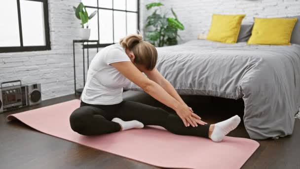 mooie jonge blonde vrouw zitten op slaapkamer vloer, opwarmen, stretching haar benen, ontspannen temidden van yoga workout - Video