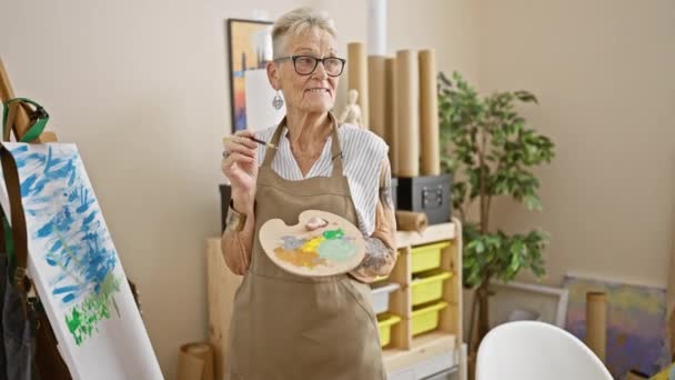 Femme artiste senior aux cheveux gris et confiante, souriant joyeusement tout en tenant son pinceau et sa palette de confiance dans le studio d'art animé. - Séquence, vidéo
