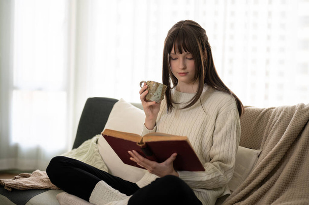 A pretty and relaxed young caucasian woman in a knitted sweater is reading a book and sipping coffee on a couch in her comfortable living room. People and leisure concepts - Photo, Image