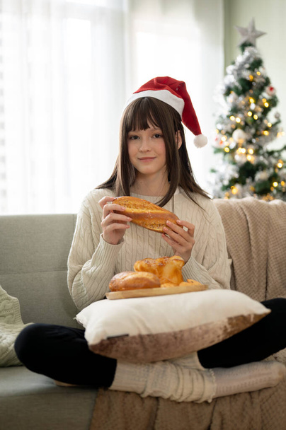 Een mooie jonge vrouw met een kerstmuts eet graag kerstdesserts op een bank in haar woonkamer en brengt haar kerstvakantie thuis door. Huiselijk leven, Kerst evenement - Foto, afbeelding