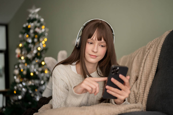 Uma mulher feliz e bonita está ouvindo música em seus fones de ouvido e usando seu smartphone enquanto estava deitada em um sofá em sua sala de estar nas férias de Natal. - Foto, Imagem