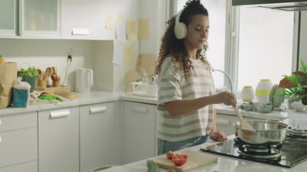Середній довгий знімок веселої дівчини в бездротових навушниках насолоджуючись музикою під час приготування їжі на кухні вдома - Кадри, відео