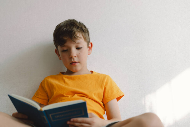 Ein süßer Junge in einem orangefarbenen T-Shirt sitzt auf einem weichen Hocker und liest ein Buch. Kopierraum. Echte Menschen und Lebensstil - Foto, Bild