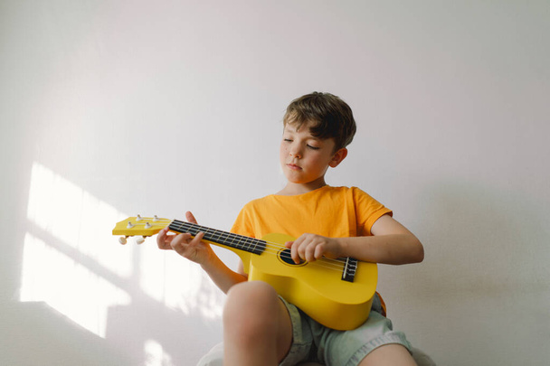 Leuke jongen leert thuis de gele ukelele gitaar spelen. Gezellig thuis. Zomervakantie levensstijl. - Foto, afbeelding
