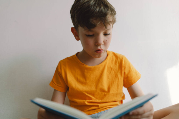 Un chico lindo con una camiseta naranja está sentado en un otomano suave leyendo un libro. Copiar espacio. Personas reales y estilo de vida - Foto, imagen