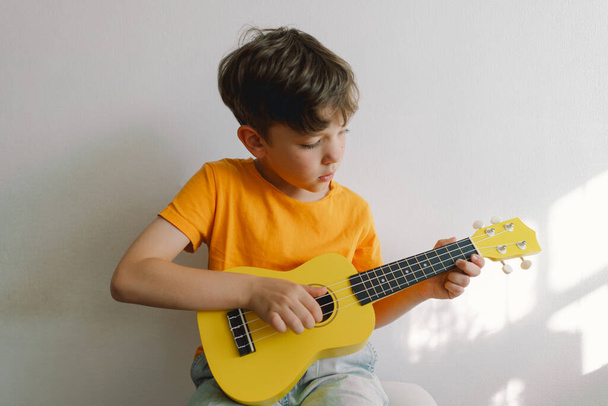 Il ragazzo carino impara a suonare la chitarra gialla Ukulele in casa. Casa accogliente. Vacanze estive stile di vita. - Foto, immagini