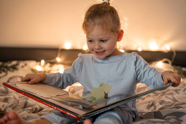 Malá holka čtení pop-up knihy v domácí posteli ve vánočním prostředí se světly pozadí. Kvalitní fotografie - Fotografie, Obrázek