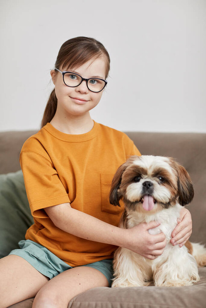 Κάθετη πορτρέτο της έφηβης με σύνδρομο Down κάθεται με χαριτωμένο σκυλί στον καναπέ και κοιτάζοντας κάμερα χαμογελώντας - Φωτογραφία, εικόνα