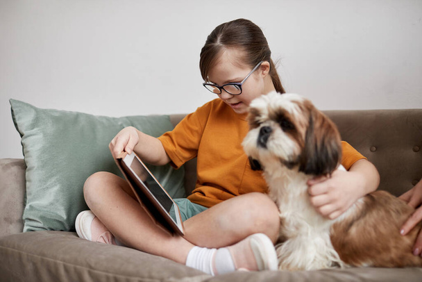 Teljes hosszúságú portré aranyos lány Down-szindróma segítségével tabletta, miközben ül a kanapén a kutya társa - Fotó, kép