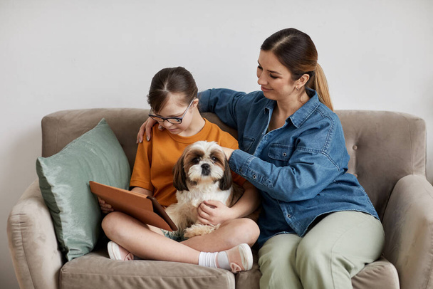Minimal portrait of caring mother and daughter with Down syndrome sitting on couch with cute dog - Photo, Image
