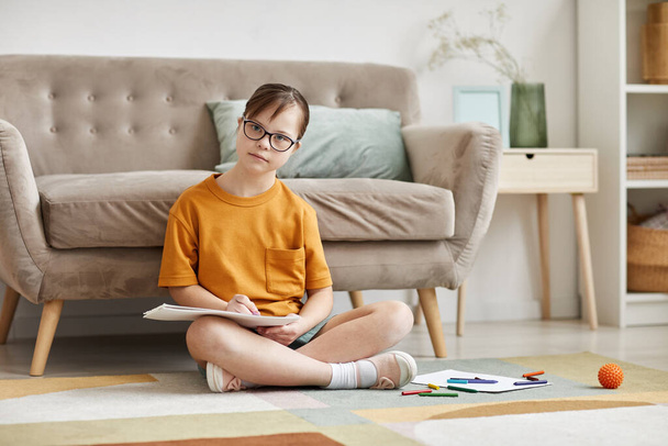 Teljes hosszúságú portré egy Down-szindrómás tinédzser lányról, aki otthon ül a padlón keresztbe tett lábbal, és a kamerába néz, másolja a helyet - Fotó, kép