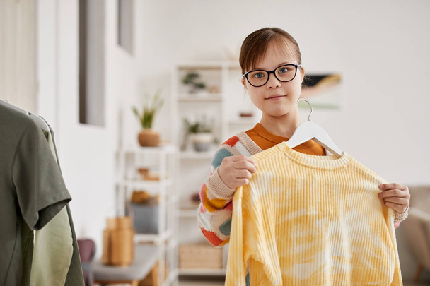 Waist up portrait of teenage girl with Down syndrome choosing clothes and looking at camera, copy space - Photo, Image